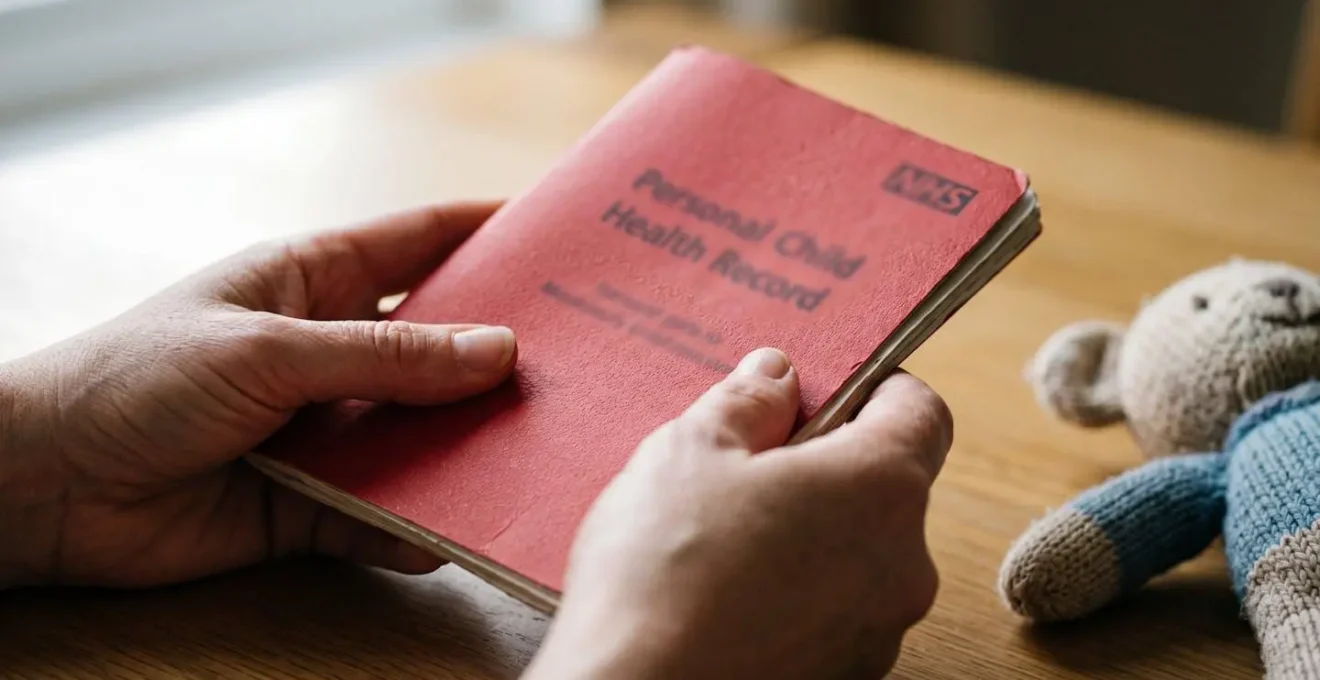 Close-up of parent's hands gently holding UK NHS Red Book with child's comfort toy nearby in soft natural light